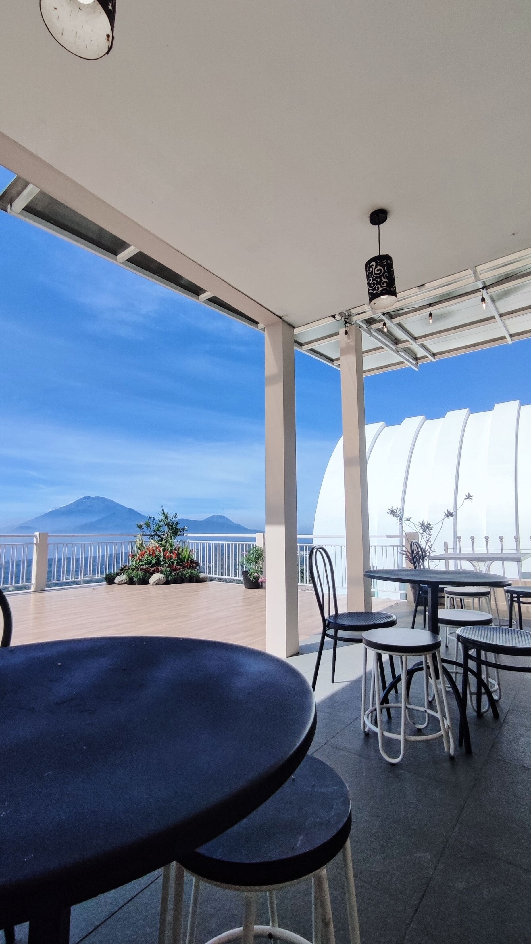 An outdoor patio with tables and chairs, a view of the sea, and a mountain in the distance.