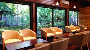 Un área de estar con sillas y mesas, ventanas amplias que ofrecen vistas a la vegetación y una estructura de madera en el exterior.