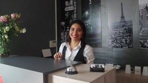 A hotel reception desk with a clock, a vase of flowers, and a cityscape poster in the background. - Metropoli Suites Ejecutivas (Puebla)