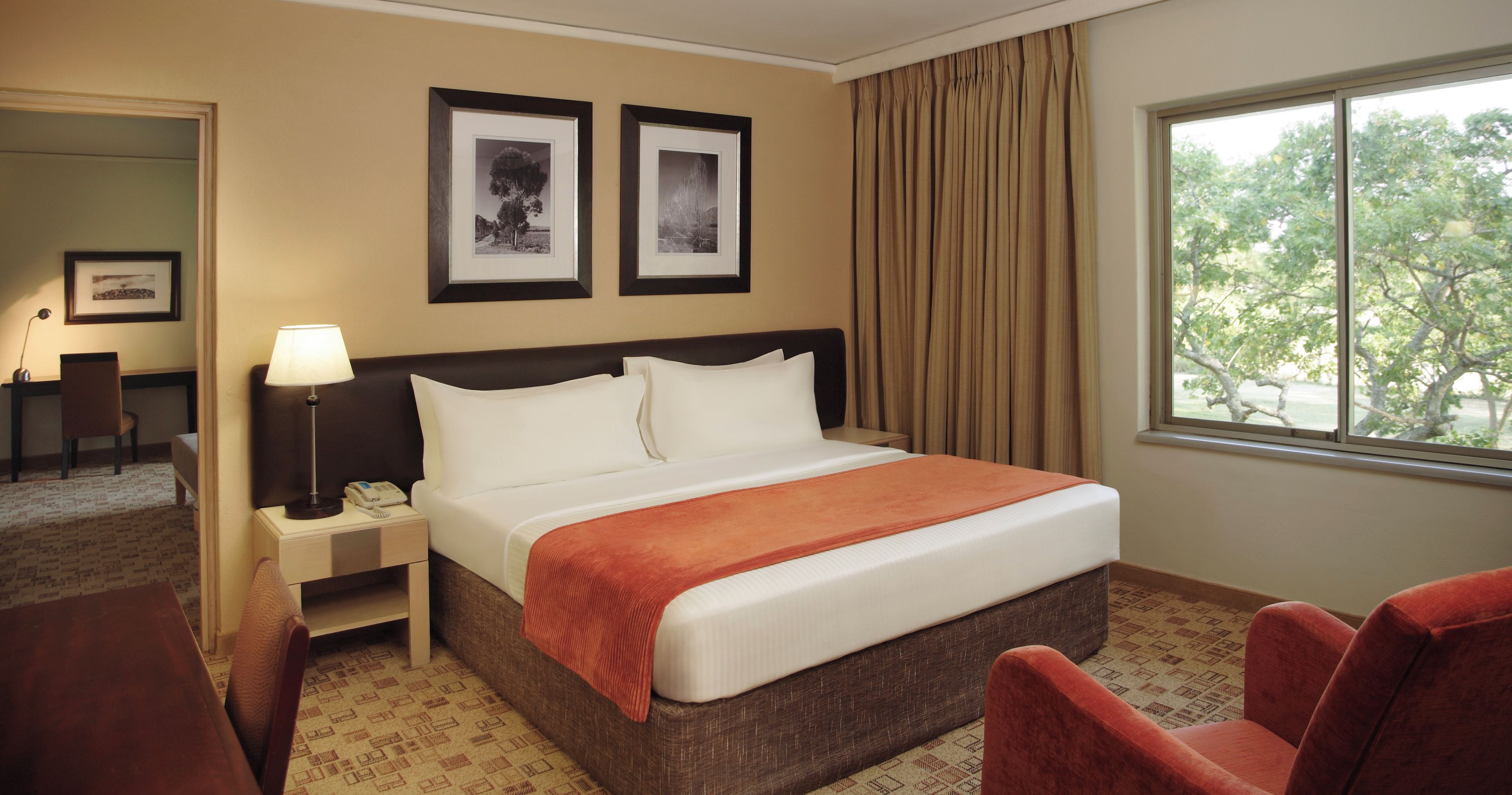 Habitación de hotel con cama, escritorio, silla, mesita de noche, lámpara y dos cuadros en la pared.