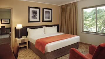 Habitación de hotel con cama, escritorio, silla, mesita de noche, lámpara y dos cuadros en la pared.