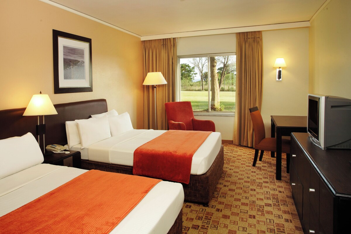 A hotel room with two beds, a desk with a television, a chair, and a window with curtains.