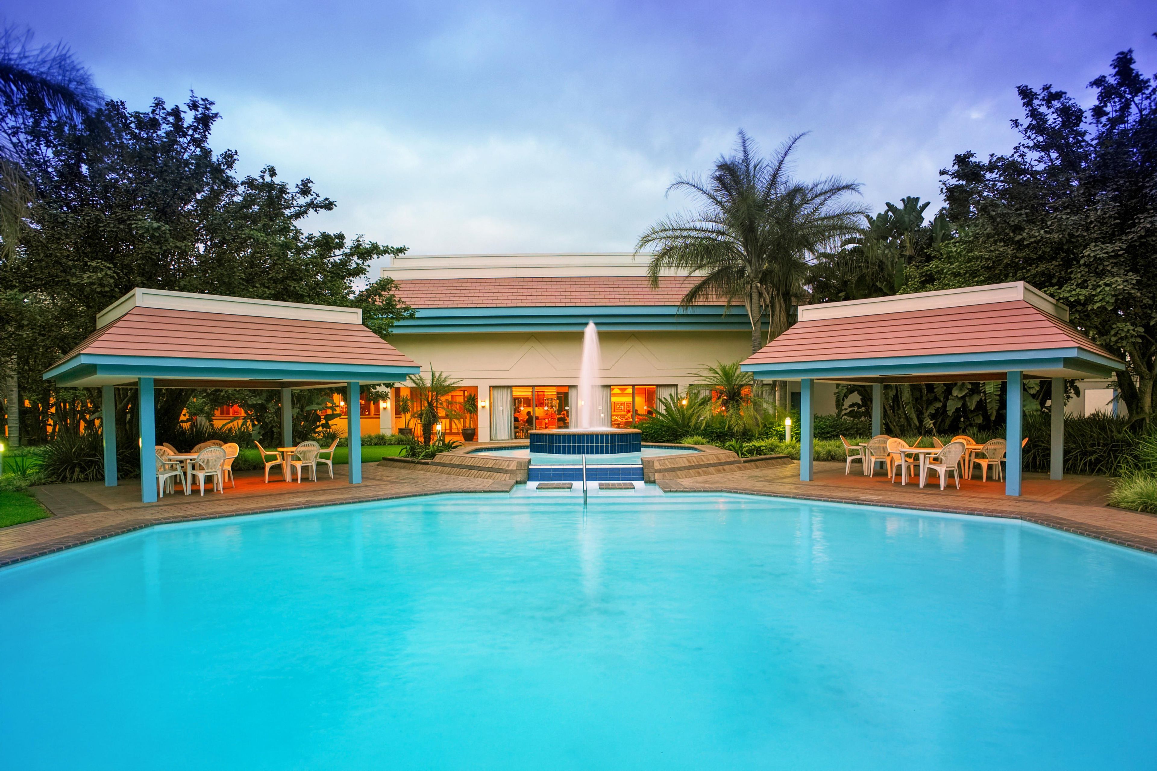 A swimming pool with a central fountain, surrounded by covered seating areas and a building with a red roof in the background.