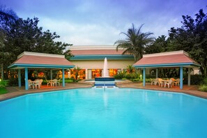A swimming pool with a central fountain, surrounded by covered seating areas and a building with a red roof in the background.