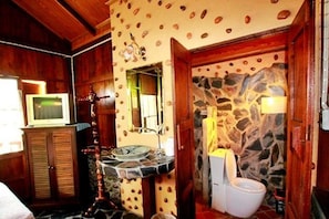 A bathroom with a stone wall, wooden cabinets, a sink with a round mirror, and a toilet. - J&J Guesthouse (Sukhothai)
