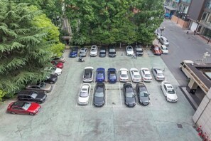 Un estacionamiento con varios coches aparcados en filas, rodeado de árboles y edificios.