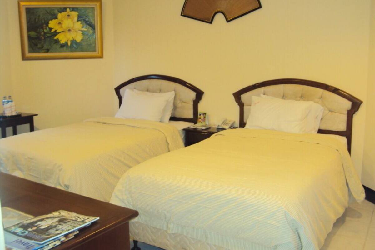 Habitació d'hotel amb dos llits individuals, una taula de nit amb una làmpada, una taula petita amb una revista i un quadre a la paret.