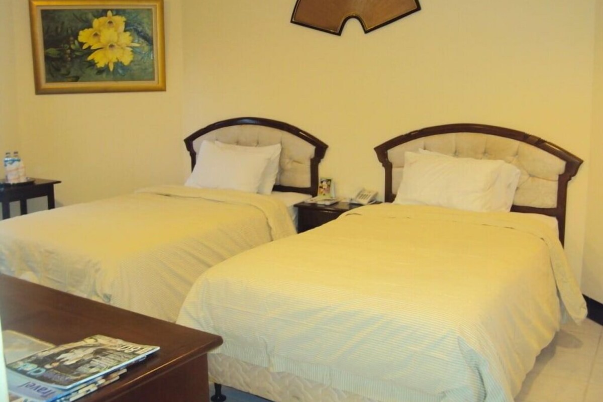 A hotel room with two beds, a bedside table with a lamp, a painting on the wall, and a small table with a magazine.