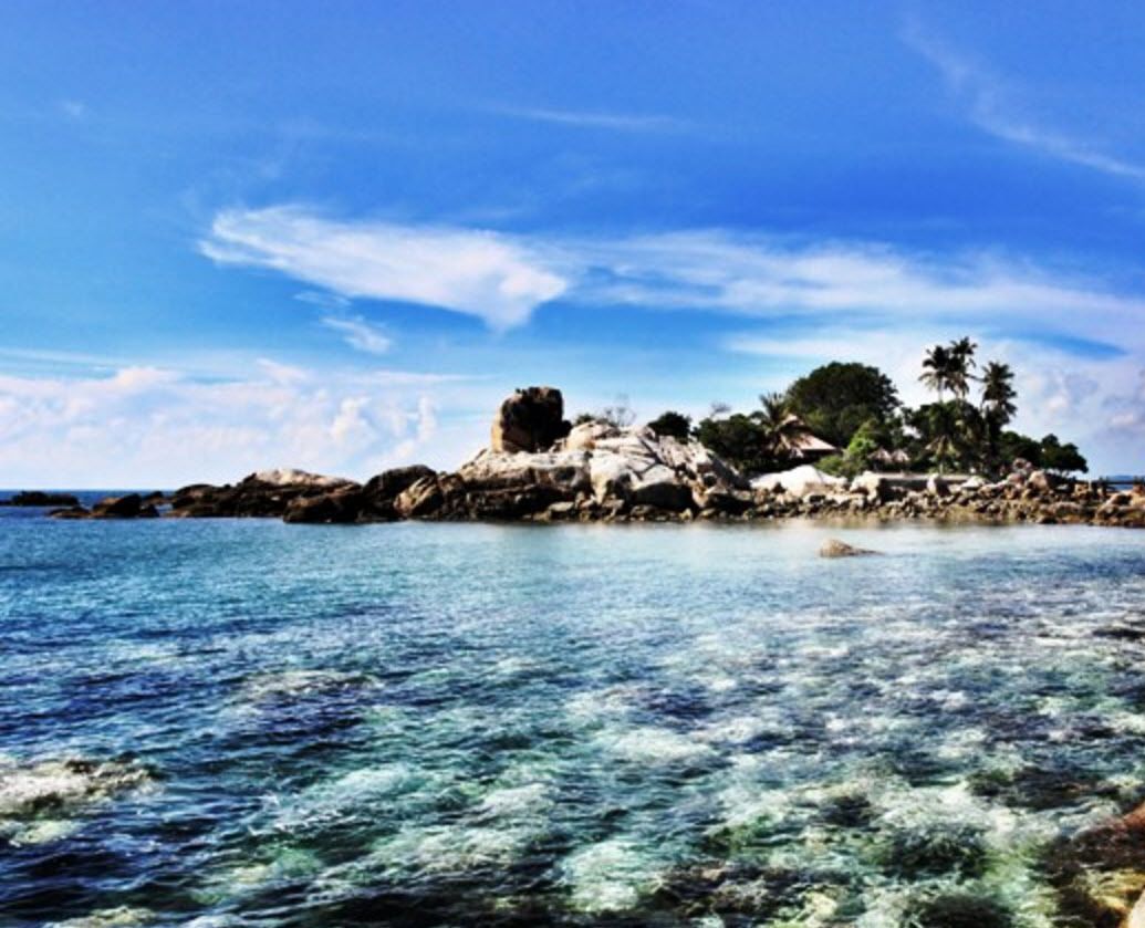A rocky coastal landscape with clear blue water and a small island with palm trees.