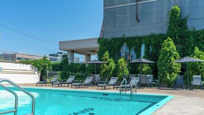 A swimming pool with lounge chairs and umbrellas, surrounded by greenery and a modern building. - Hôtel Lindbergh par JARO (Québec City)
