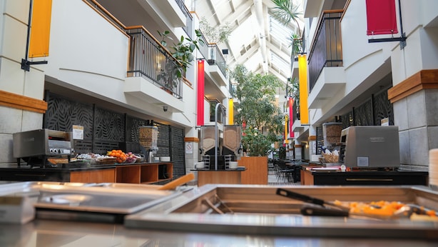 A modern hotel atrium with a food service area, including a self-service station with various food items and a coffee machine. - Hôtel Lindbergh par JARO (Québec City)