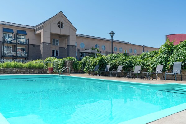 A swimming pool with lounge chairs and a building with multiple windows and balconies in the background. - Hôtel Lindbergh par JARO (Québec City)