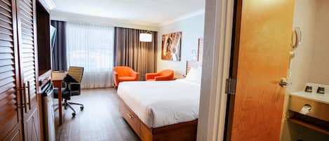 A hotel room with a bed, two orange chairs, a desk with a chair, a window with curtains, and a painting on the wall.