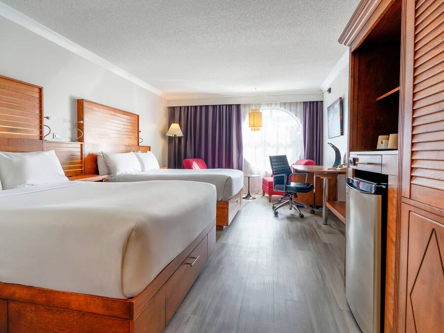 Habitación de hotel con dos camas, un escritorio, una silla, un refrigerador y un ventanal con cortinas.
