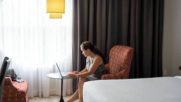A hotel room with a bed, a seating area with a laptop, and a window with curtains.