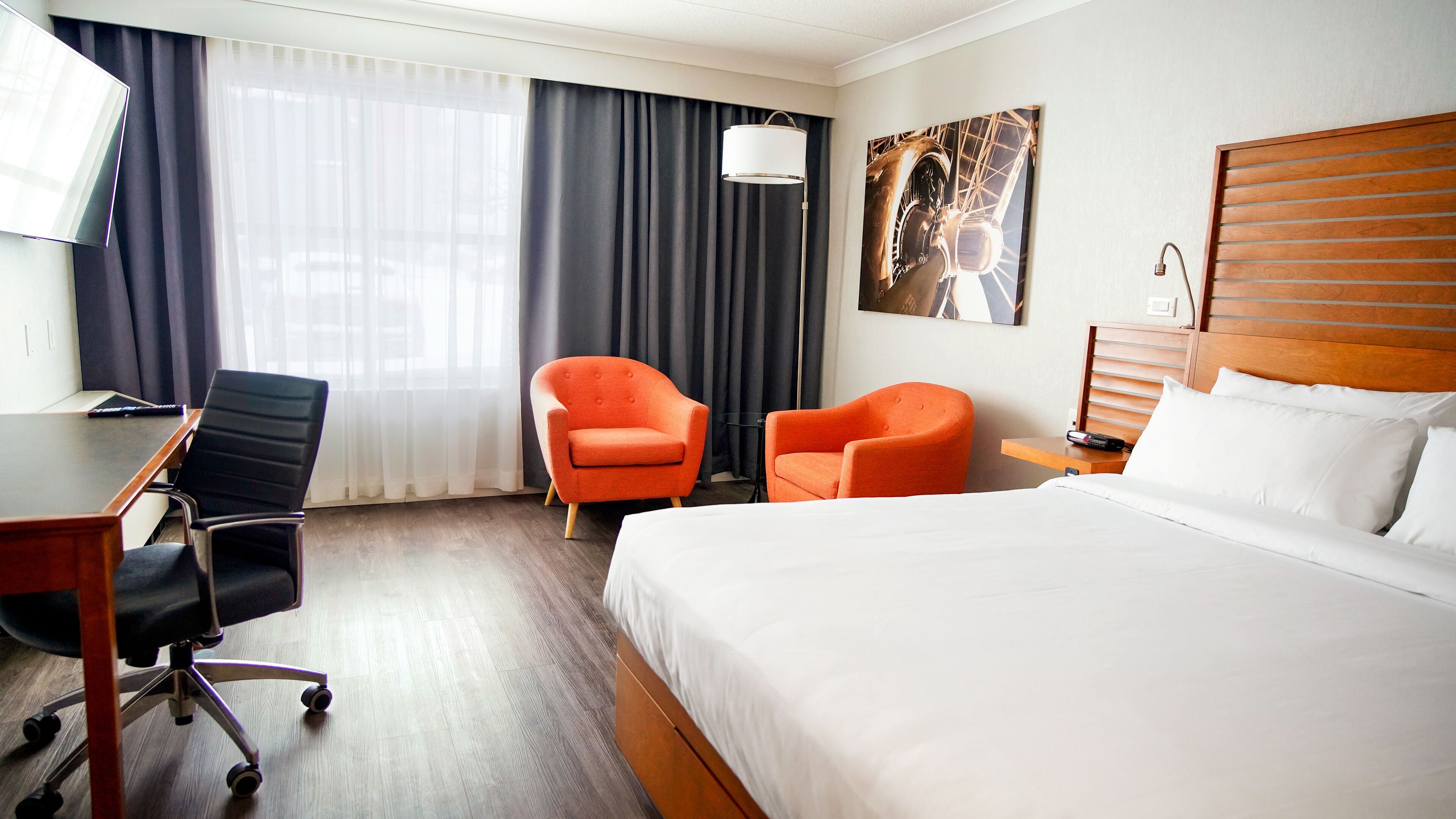 Una habitación de hotel con una cama, dos sillones naranjas, un escritorio con una silla, una lámpara y un cuadro en la pared.