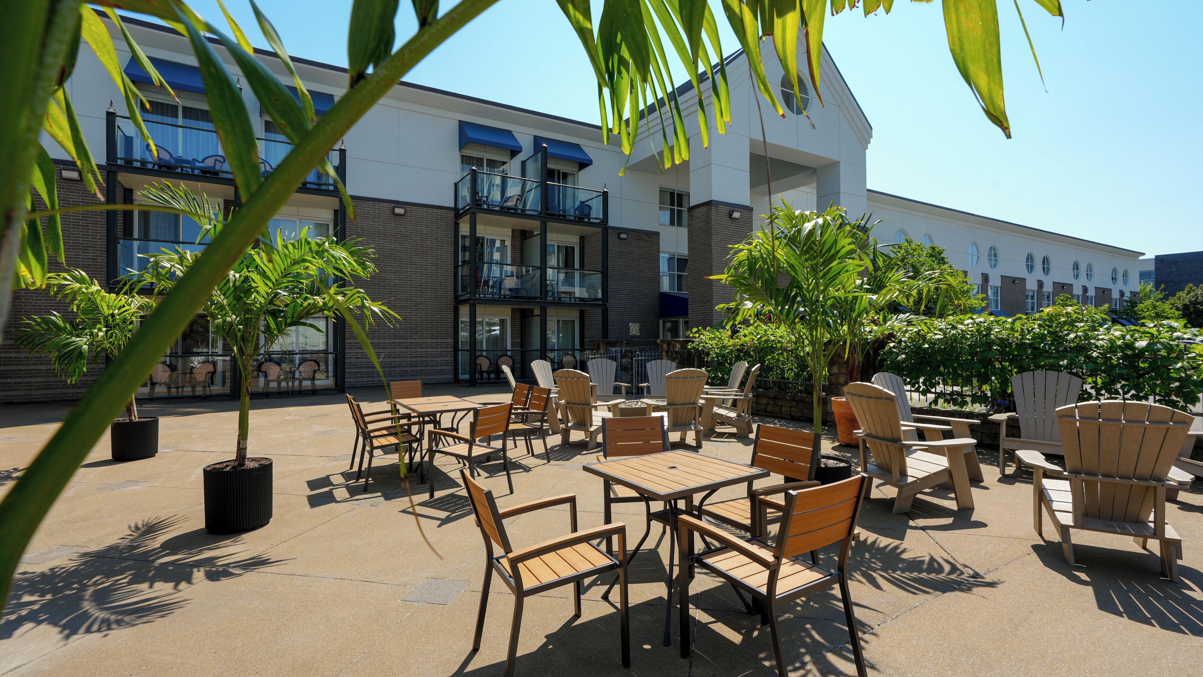 An outdoor seating area with tables and chairs, surrounded by potted plants and overlooking a building with multiple balconies.