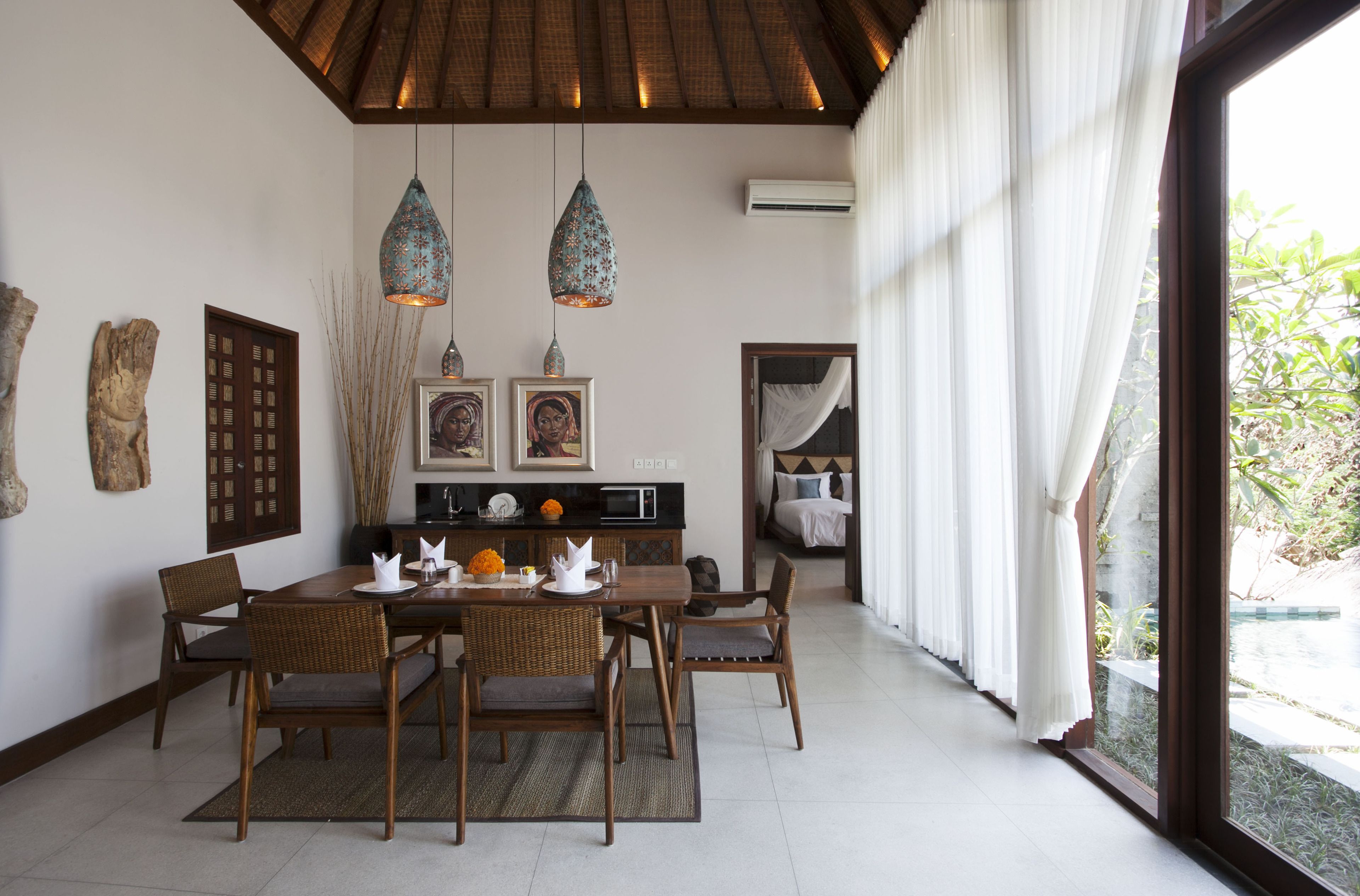 A dining area with a wooden table and chairs, two hanging pendant lights, and a view of the outdoors through a large window.