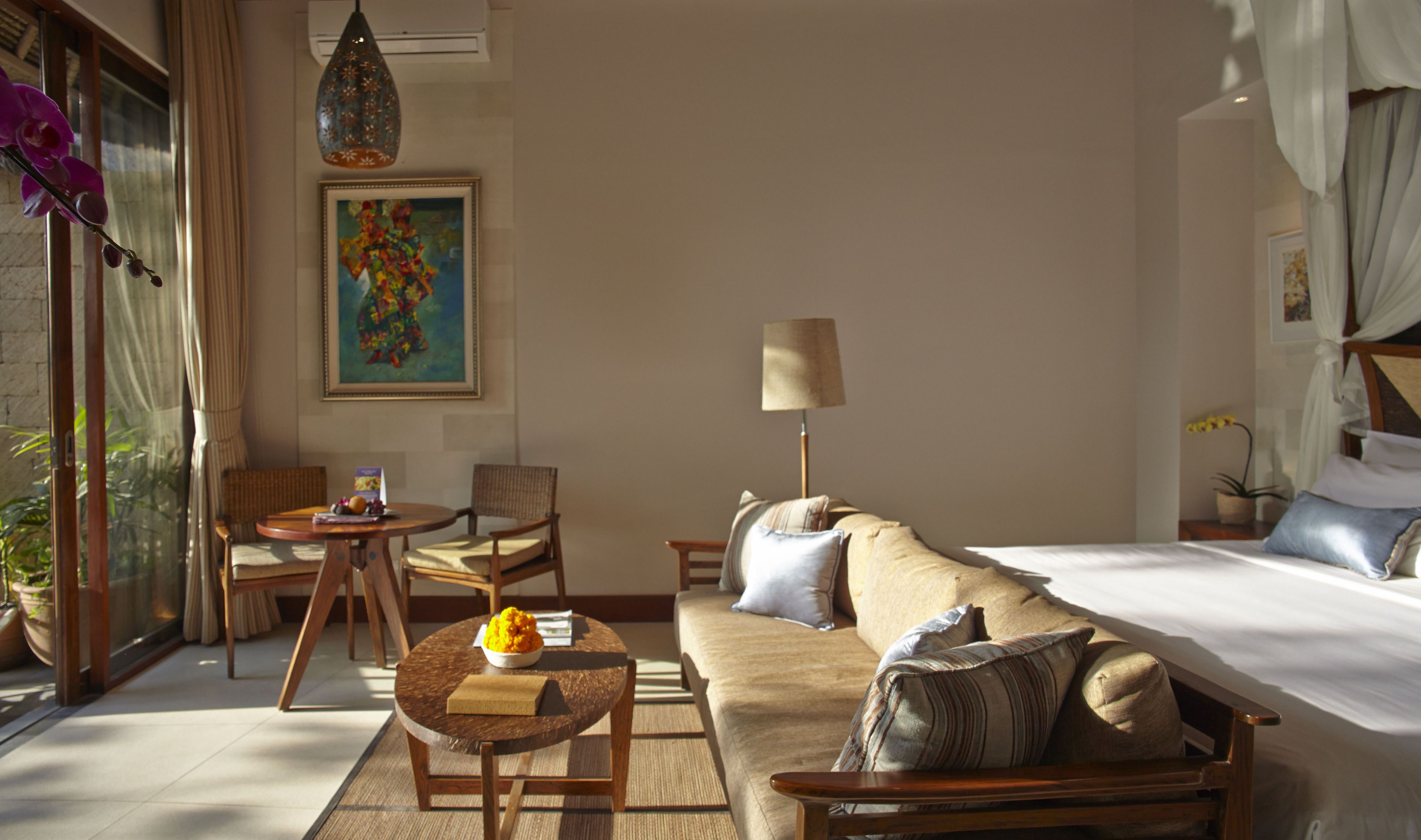 A hotel room with a bed, sofa, chairs, a small table with a bowl of fruit, a lamp, and a painting on the wall.