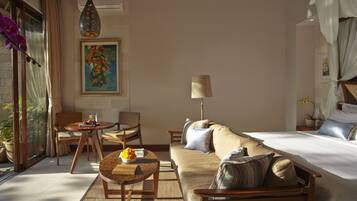 A hotel room with a bed, sofa, chairs, a small table with a bowl of fruit, a lamp, and a painting on the wall.