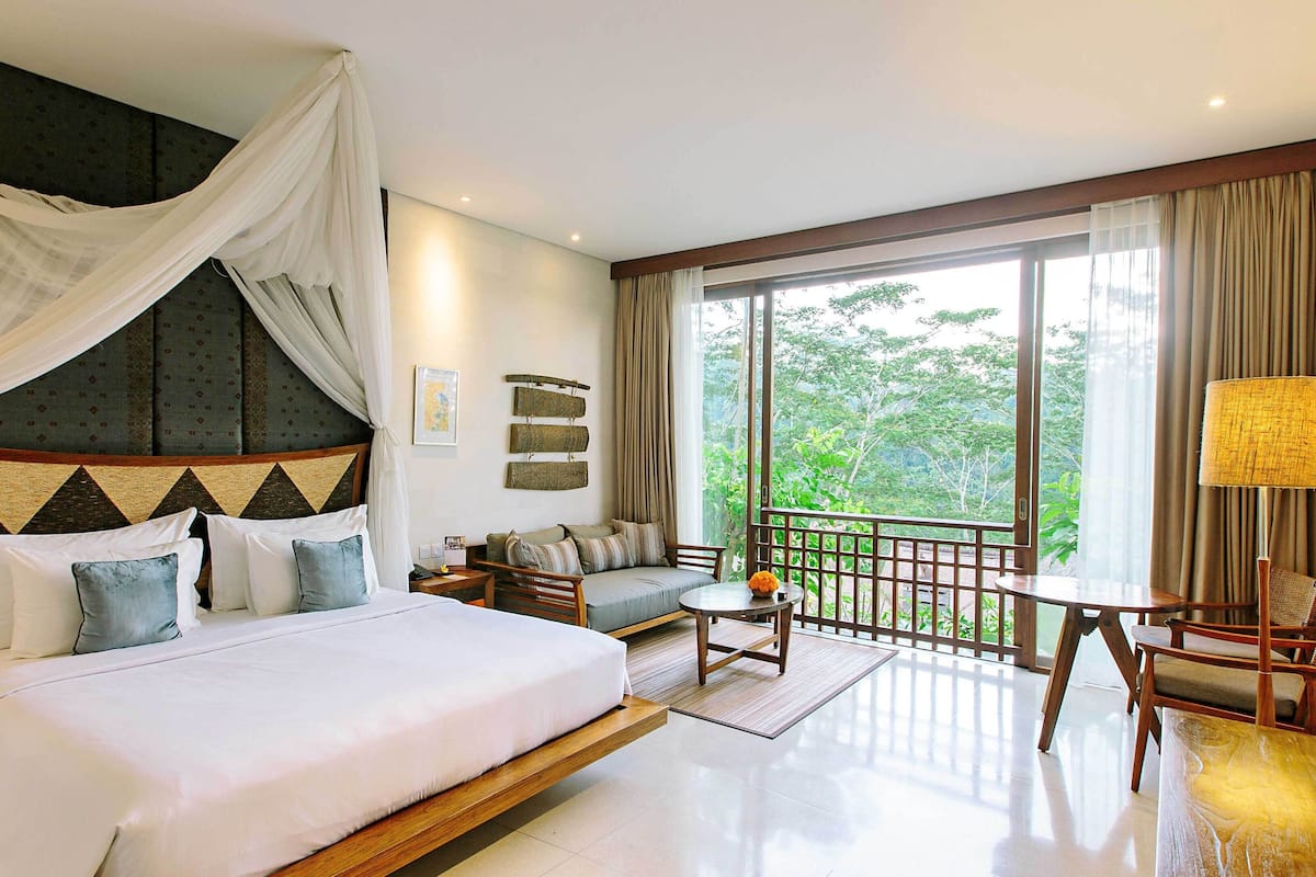 A bedroom with a large bed, a sofa, a balcony, and a view of greenery.
