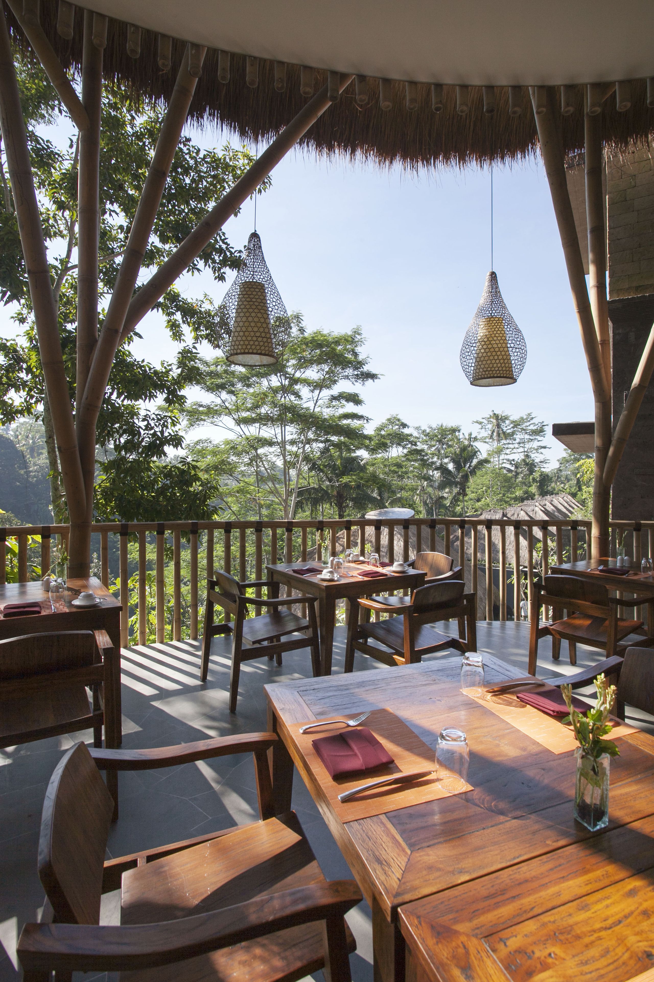 An outdoor dining area with wooden tables and chairs, thatched roof, hanging lamps, and a view of lush greenery.