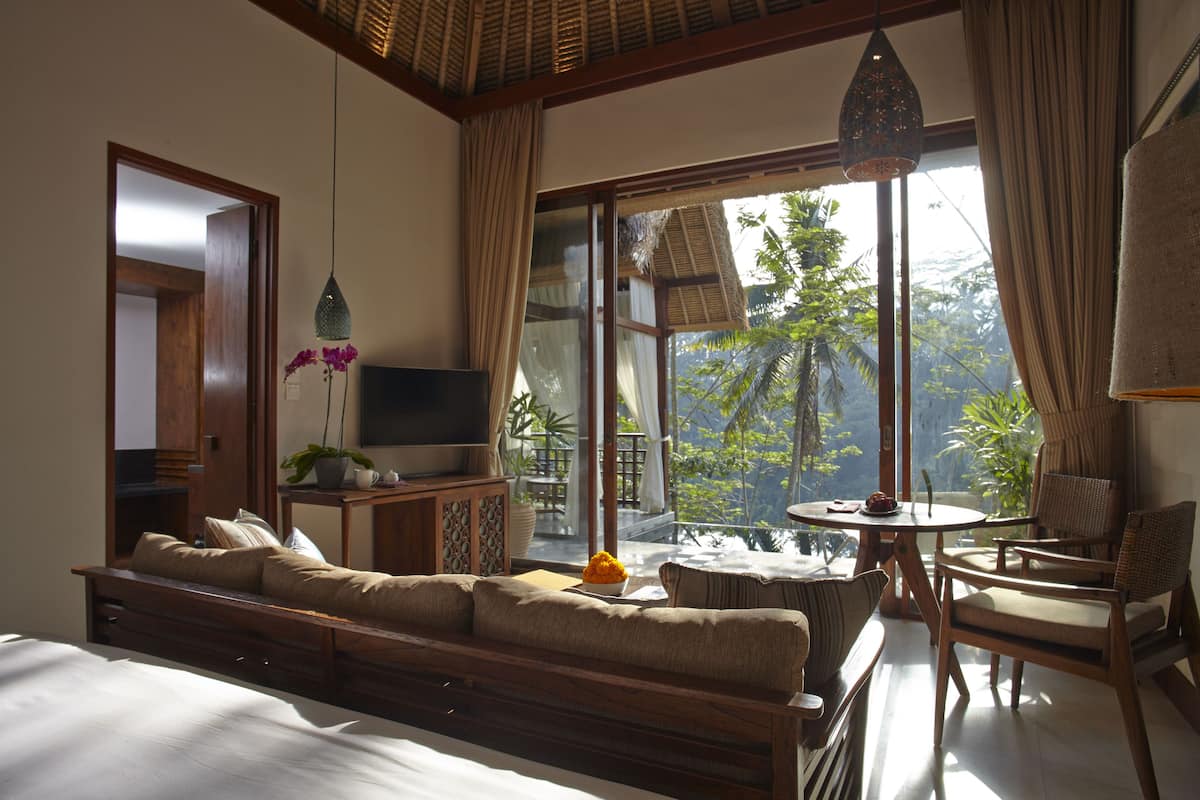 A bedroom with a large bed, a sofa, a TV, a small table, and a window with a view of greenery.
