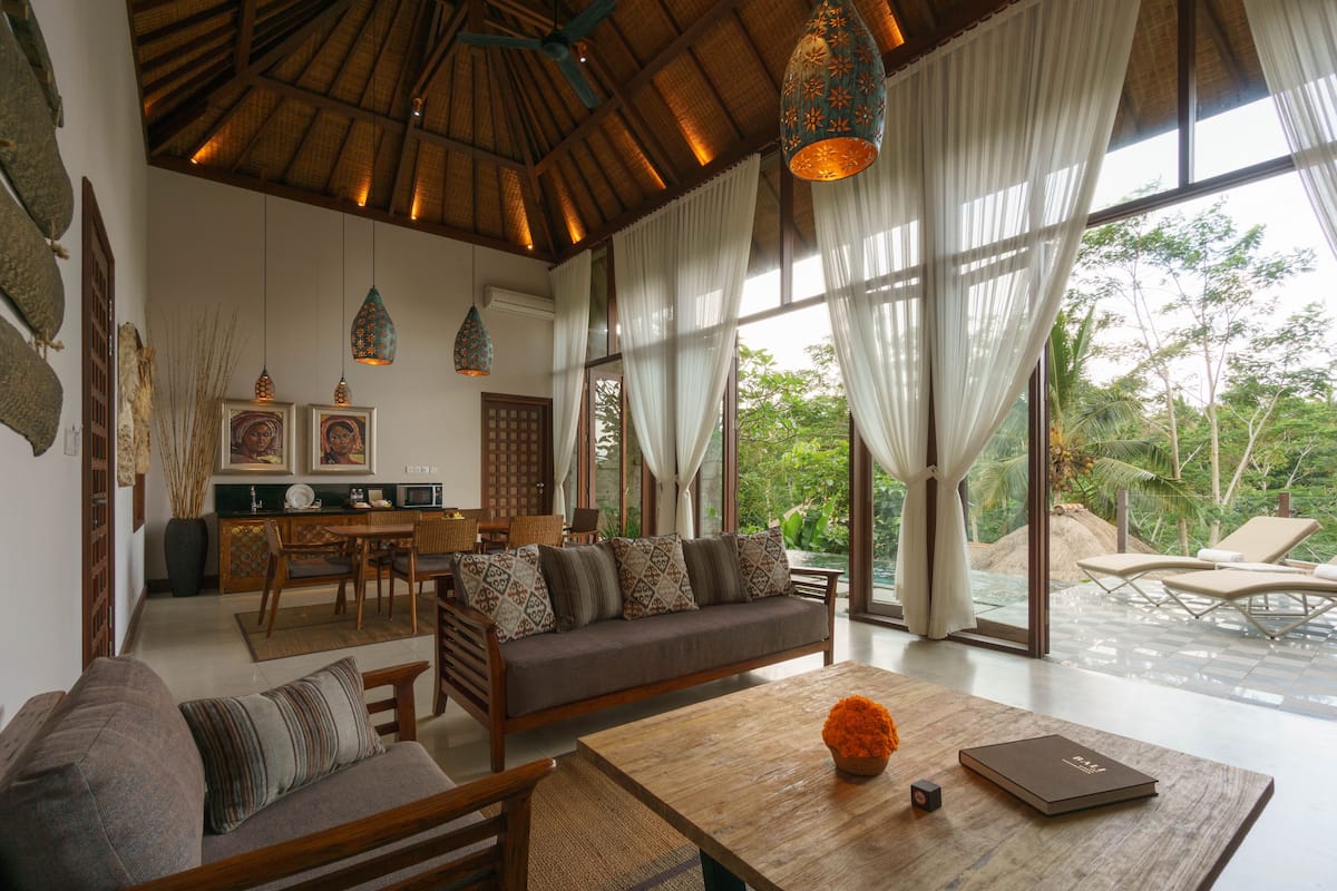 A spacious living area with a sofa, dining table, and chairs, large windows with sheer curtains, and hanging pendant lights.