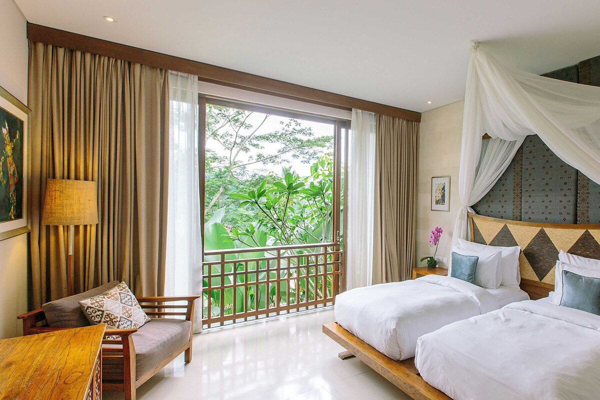 A hotel room with two beds, a balcony with a view of greenery, and a large window with curtains.