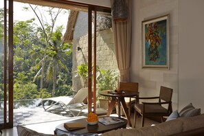 A room with a large window overlooking a tropical garden, a wicker table with a bowl of fruit, and a painting on the wall. - Tejaprana Resort & Spa (Ubud)