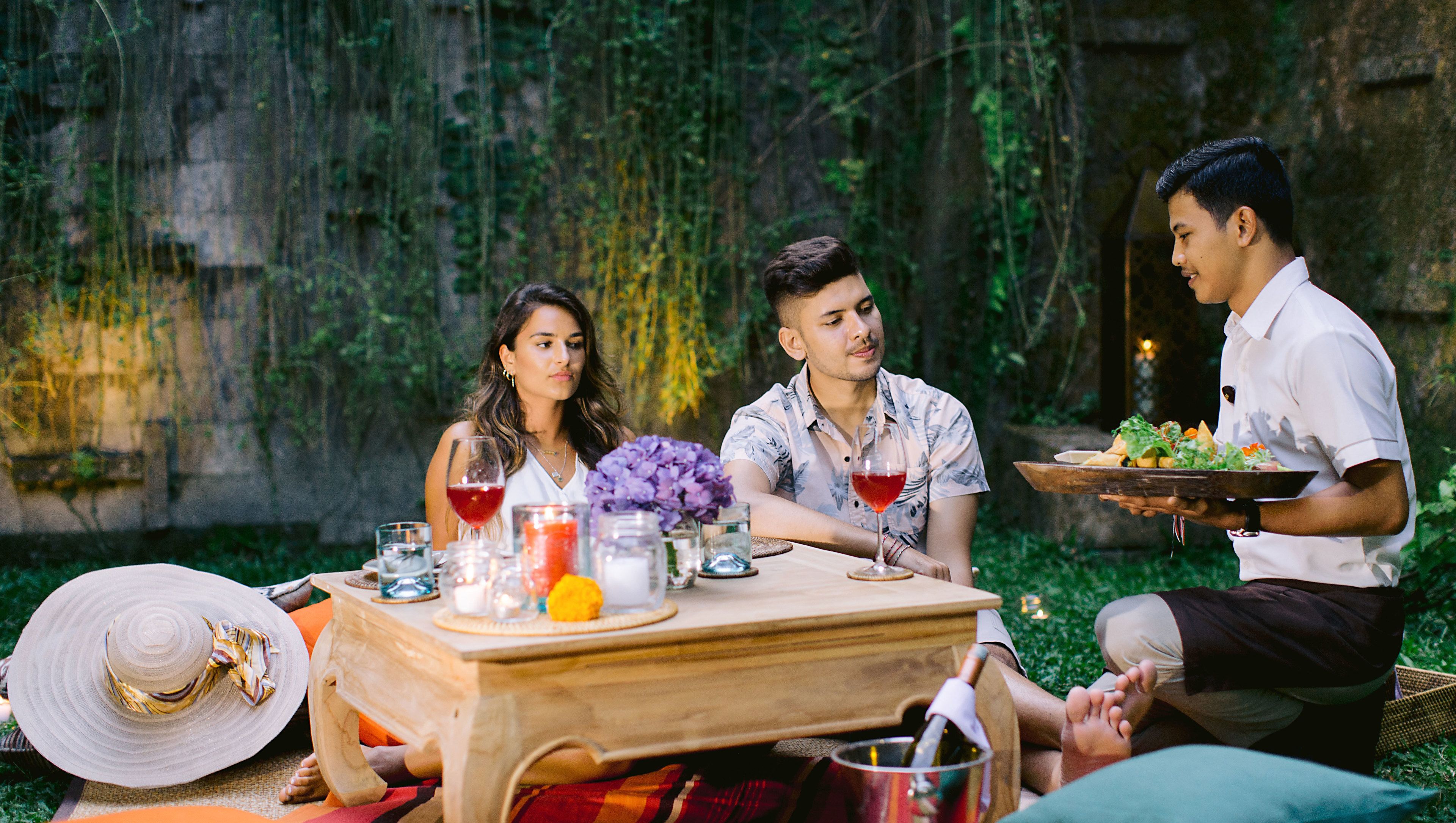 A group of people dining outdoors with a table set for a meal, surrounded by greenery and a stone wall.