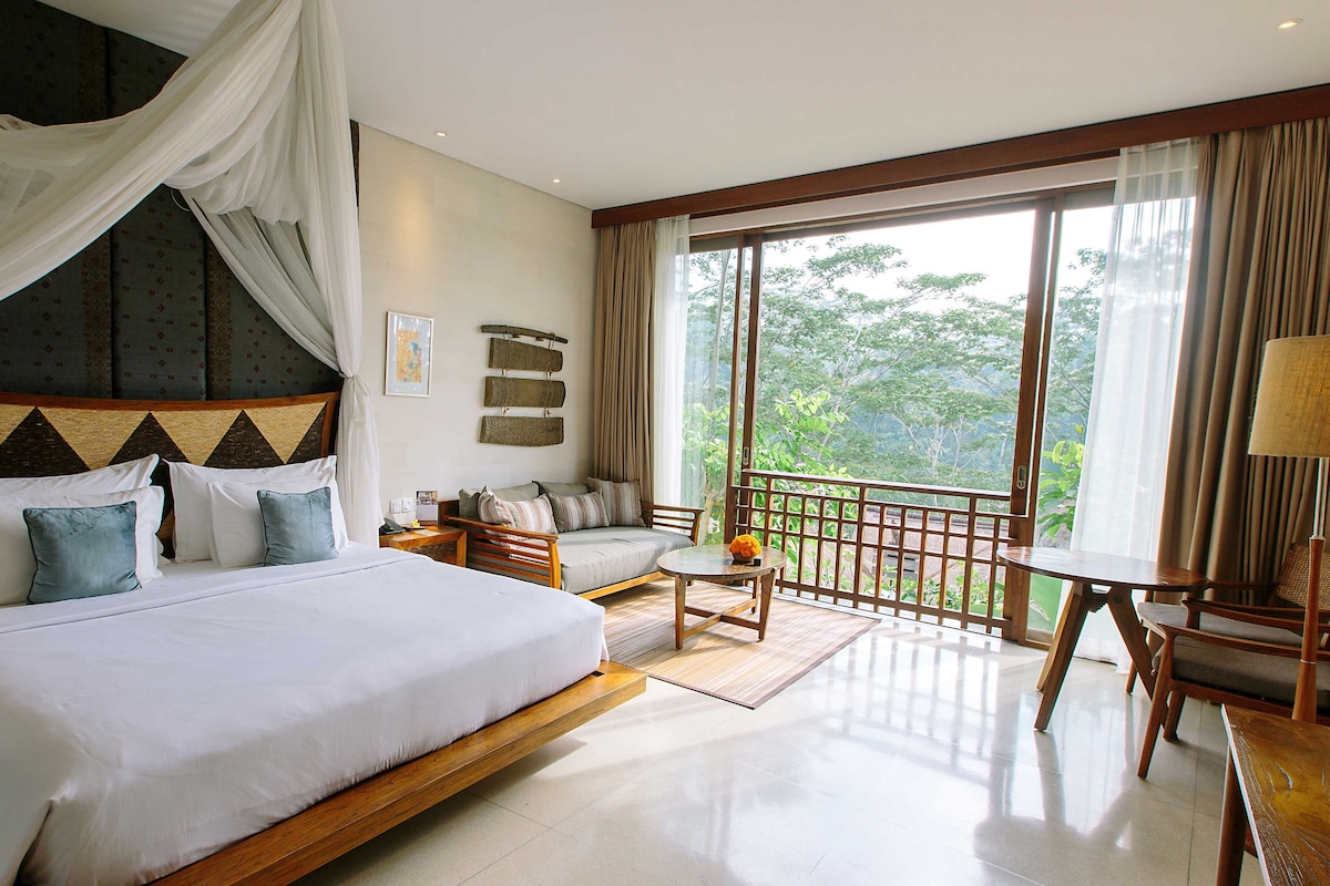 A bedroom with a large bed, a sofa, a small table, and a balcony with a view of greenery.