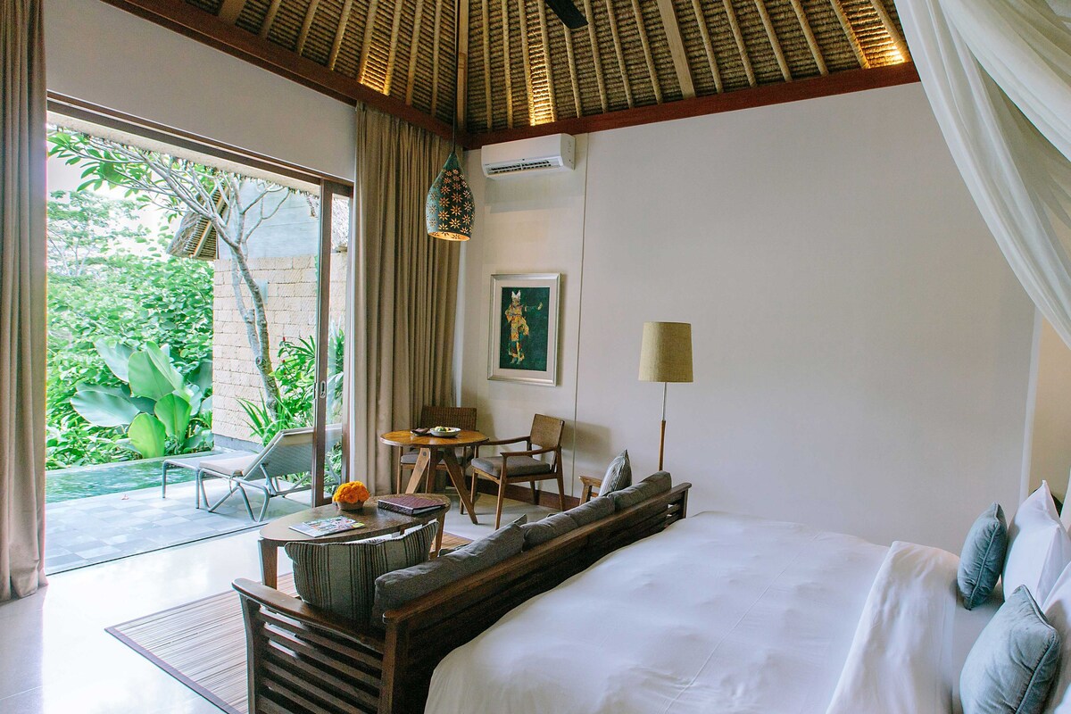 A bedroom with a large bed, a desk with a chair, a table, and a view of greenery outside.