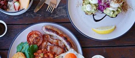 A table with three plates of food, including a plate with sausages, beans, and a fried egg, another with avocado and vegetables, and a third with a salad and a lemon wedge.
