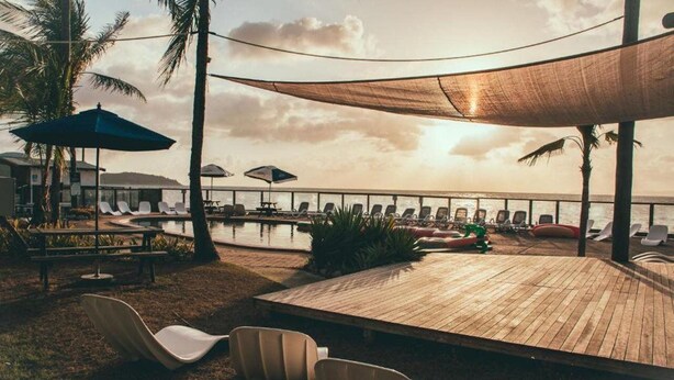 A wooden deck with lounge chairs and umbrellas, overlooking a pool and ocean view. - Base Backpackers Magnetic Island - Hostel (Nelly Bay)