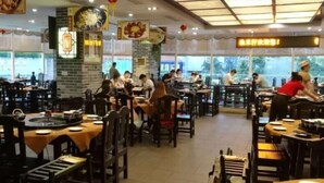 A restaurant with multiple tables and chairs, some with round tables and others with rectangular ones. There are signs hanging from the ceiling and a brick wall in the background. - Hongfeng Hotel Shenzhen (SHENZHEN)