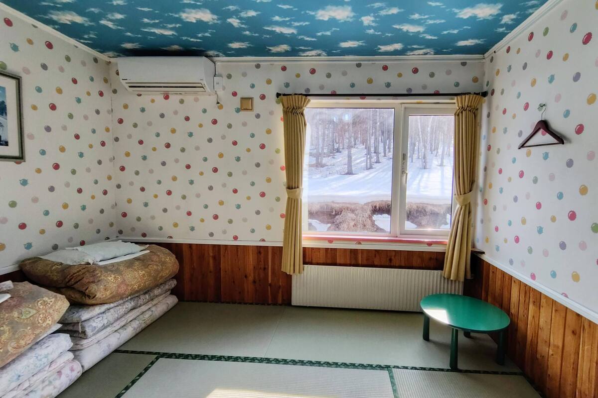 Een kamer met stippenbehang, een bed met beddengoed met dessin, een raam met gordijnen, een radiator en een klein tafeltje.