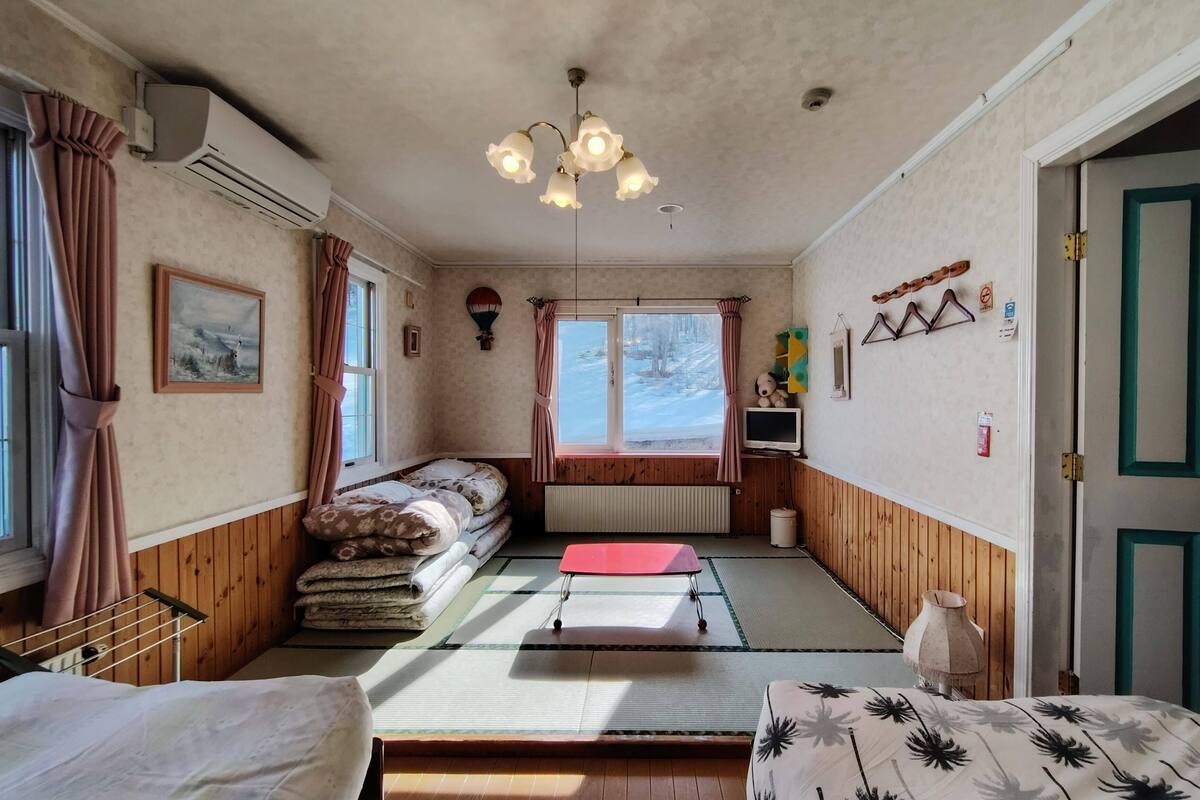 Een kamer met een bed, een klein tafeltje, een stoel, een verwarming, een raam met gordijnen, een wandairconditioner, een plafondlamp en een deur.