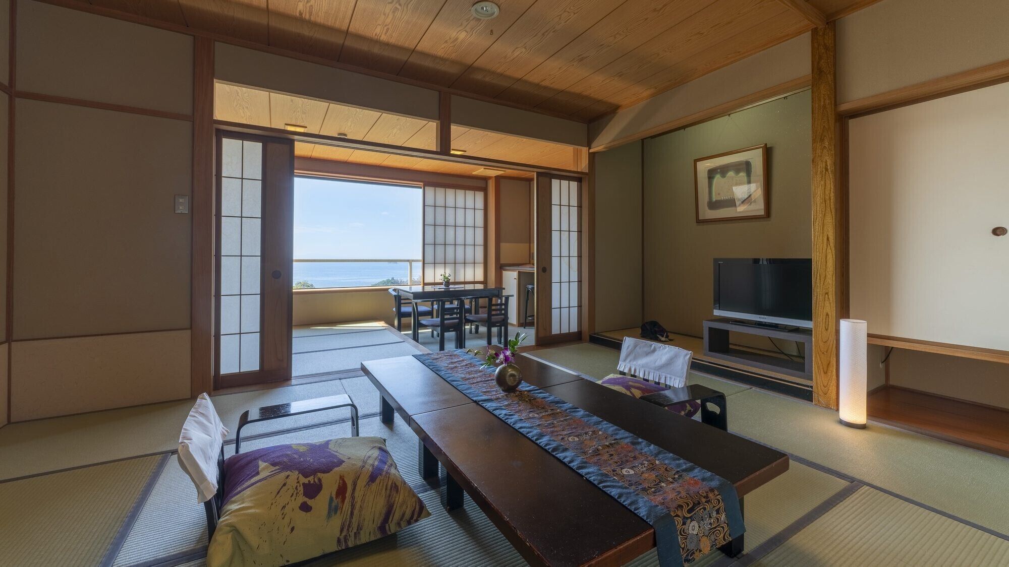A traditional Japanese room with tatami flooring, a low table with a patterned runner, a television, and a view of the sea.