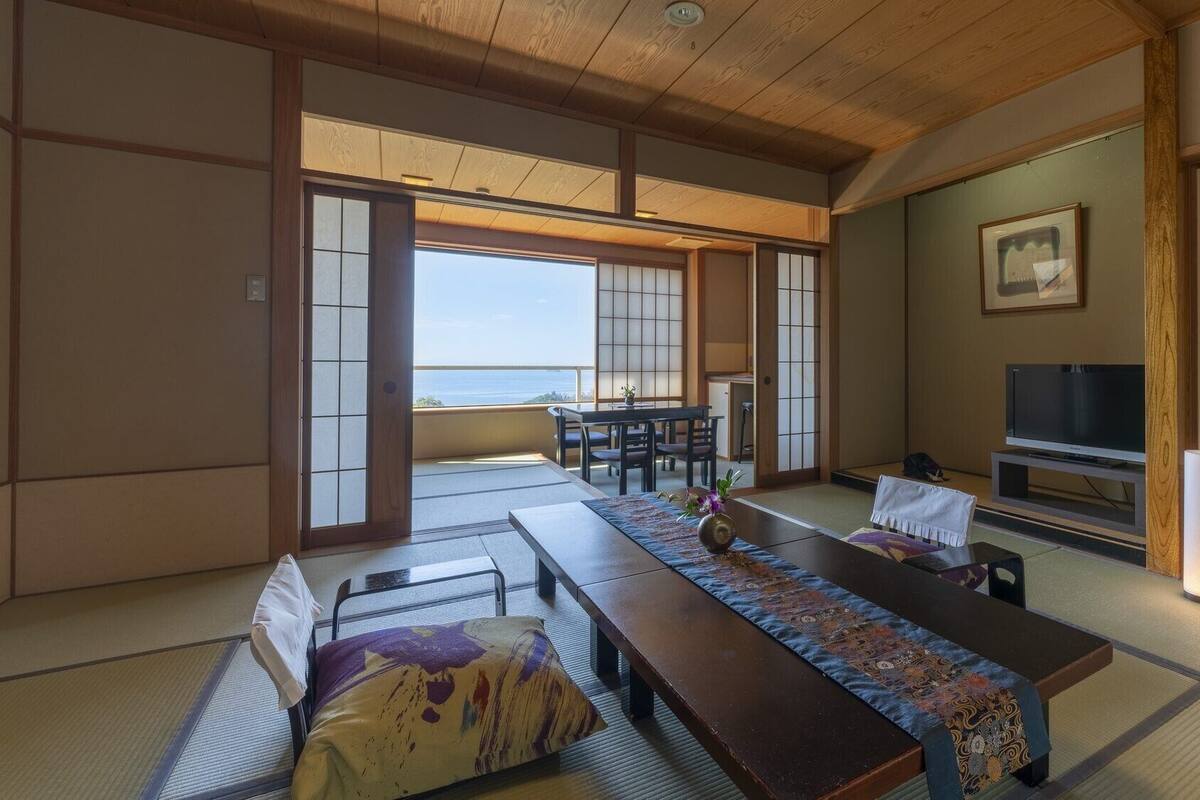 A traditional Japanese room with tatami flooring, a low table with a patterned runner, a television, and a view of the sea.