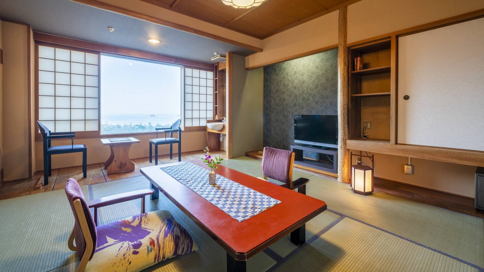 A traditional Japanese-style room with tatami flooring, a low table with a patterned mat, a television, and a view through a shoji screen.