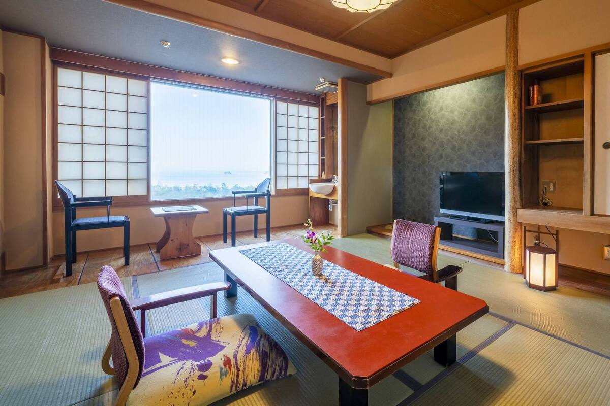 Ein traditioneller japanischer Raum mit Tatami-Boden, einem niedrigen Tisch mit gemustertem Untersetzer, einem Fernseher und Blick durch einen Shoji-Bildschirm.