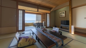 A traditional Japanese room with tatami flooring, a low table with a patterned runner, a sofa, a television, and a sliding door leading to a balcony with a view of the sea. - Hiranoya (Gamagori)