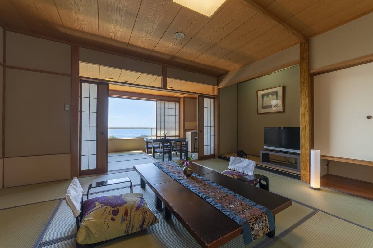 A traditional Japanese room with tatami flooring, a low table with a patterned runner, a sofa, a television, and a sliding door leading to a balcony with a view of the sea.