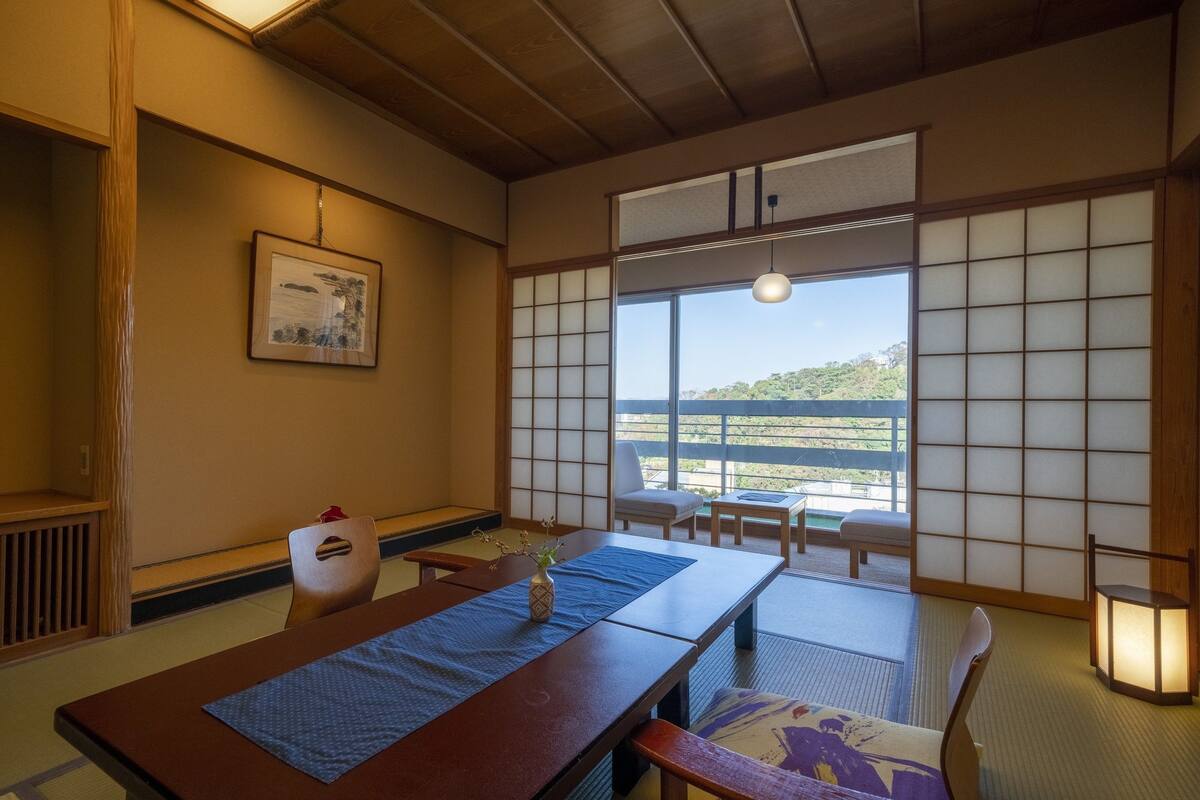 A traditional Japanese room with tatami flooring, a low table with a vase, a framed picture on the wall, and a view of a hillside through a shoji screen.