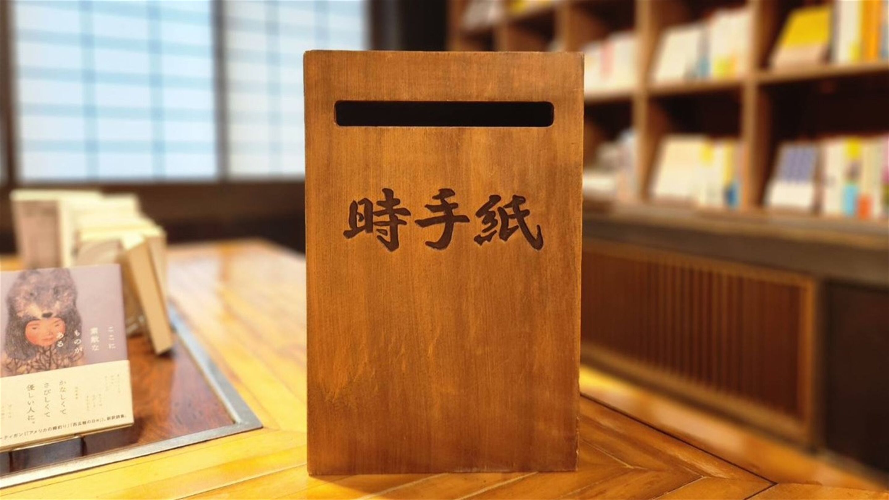 A wooden display stand with Japanese characters, placed on a wooden floor in a room with bookshelves in the background.