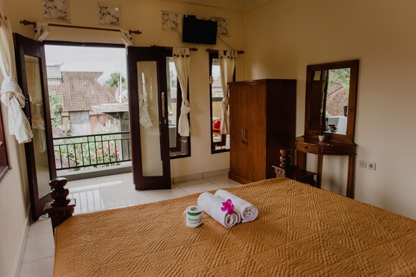 A hotel room with a bed, a wooden wardrobe, a small table with a vase, a mirror, and a balcony with a view of buildings and greenery.