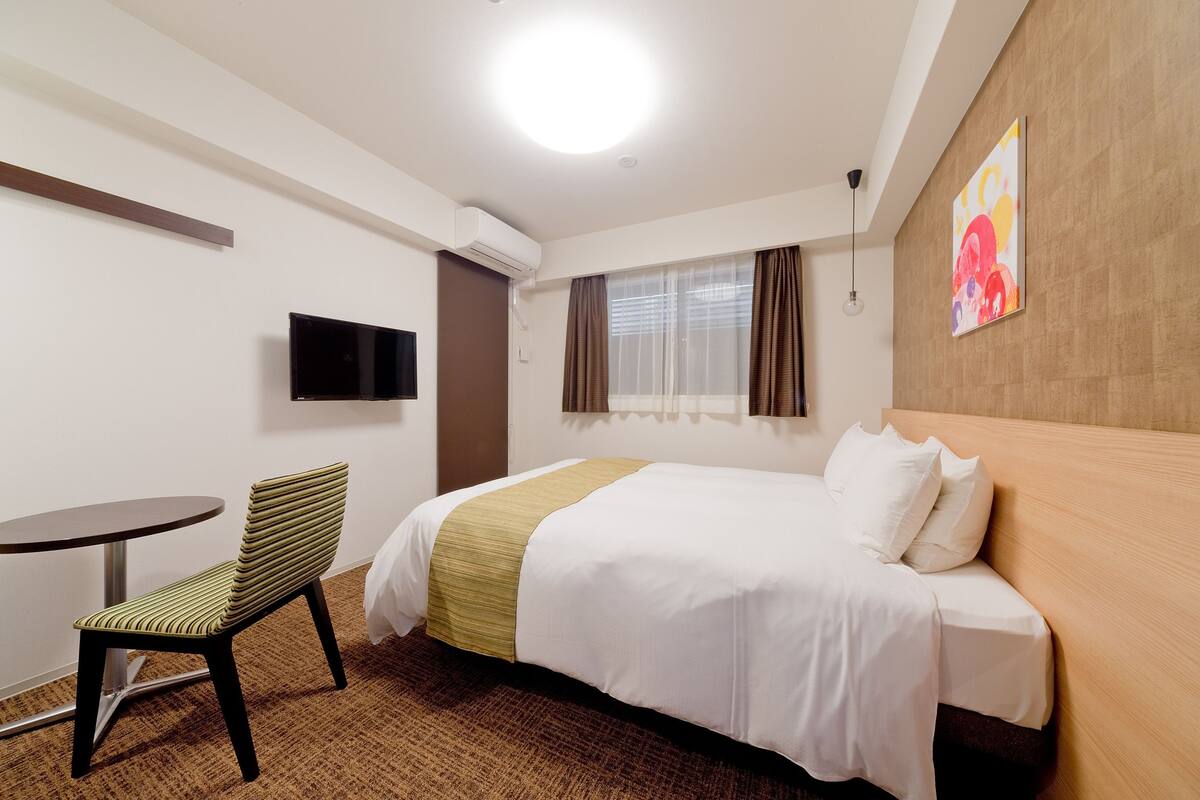 A hotel room with a bed, a TV mounted on the wall, a small round table, and a chair.