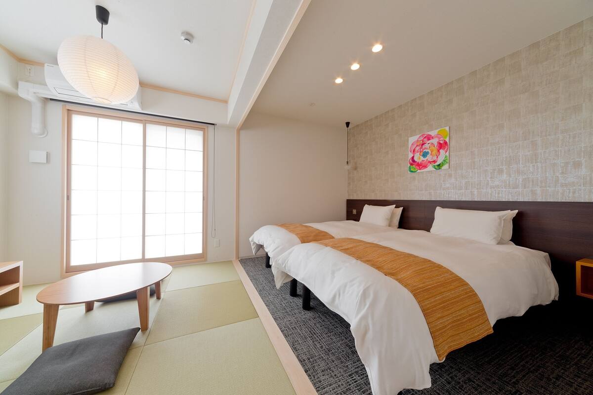 A hotel room with a large bed, a small table, tatami mats, a wall-mounted artwork, and a sliding door to the outside.