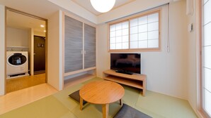 A compact living space with a tatami mat floor, a round wooden table, a television on a wooden stand, sliding doors, and a washing machine in the corner. - M’s Inn Higashiyama (Kyoto)